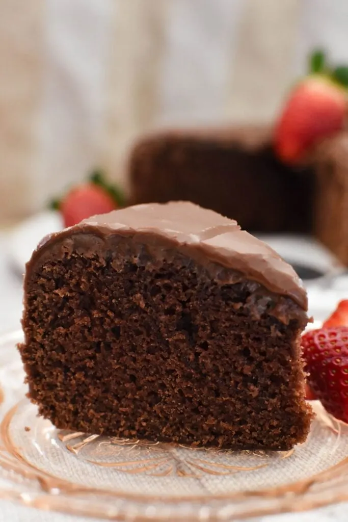 a slice of boiled chocolate cake on a plate with whole cake in background.
