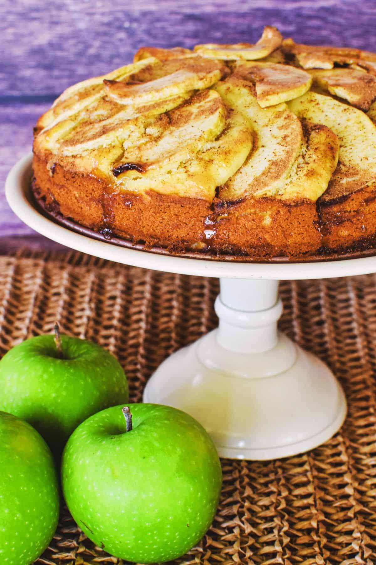 apple tea cake on cake stand with green apples in foreground.